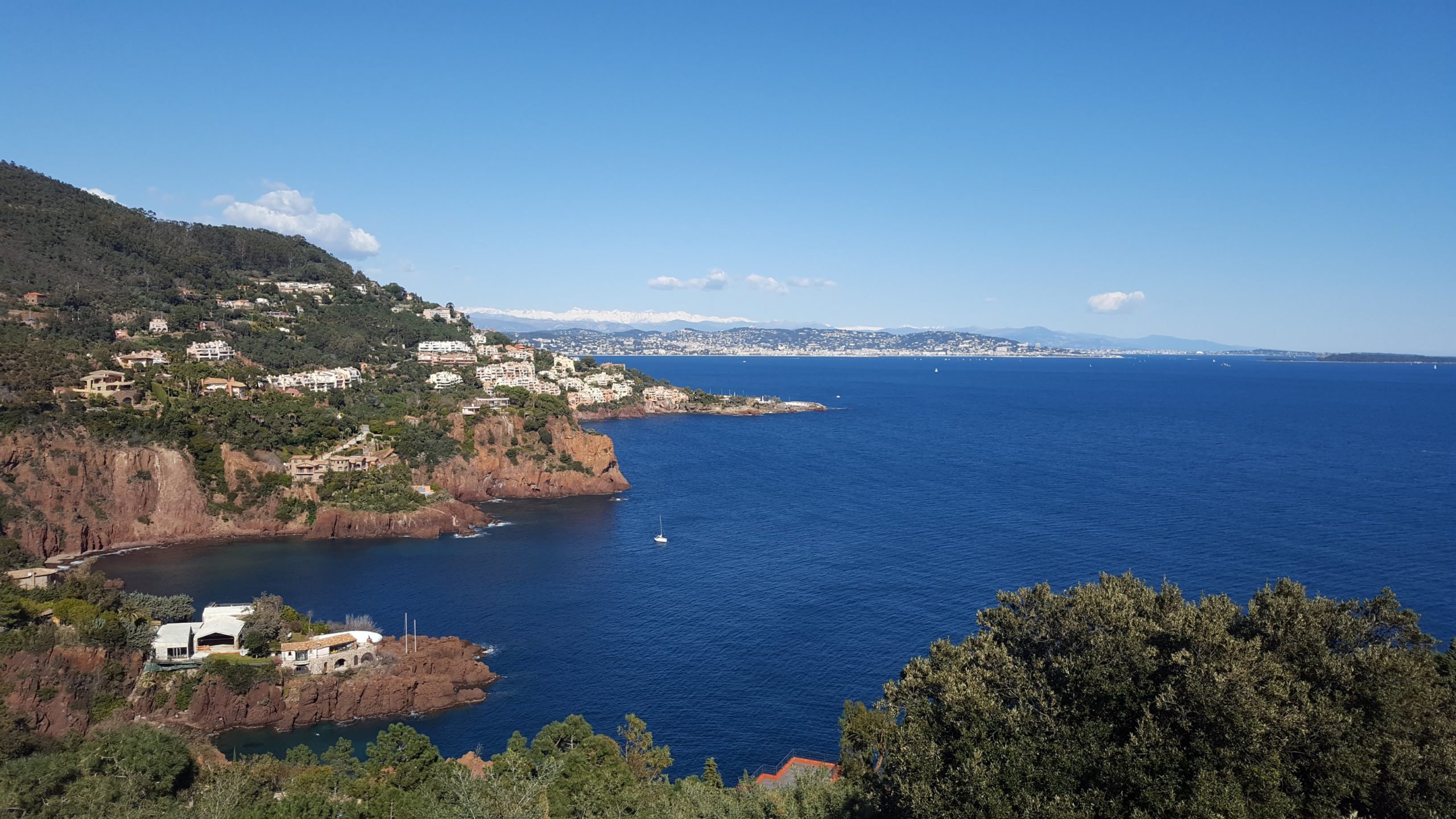 une des vues de la route de la corniche d'or permettant d'admirer les roches rouges, maisons blanches, la vegetation, la mer bleue marine et cannes et les collines au loin